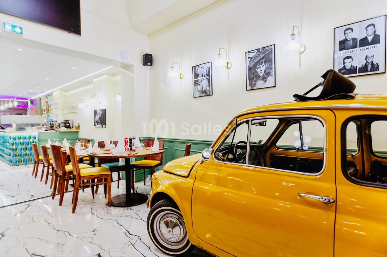 Voiture vintage jaune exposée dans un restaurant lumineux avec tables dressées et décoration murale rétro.