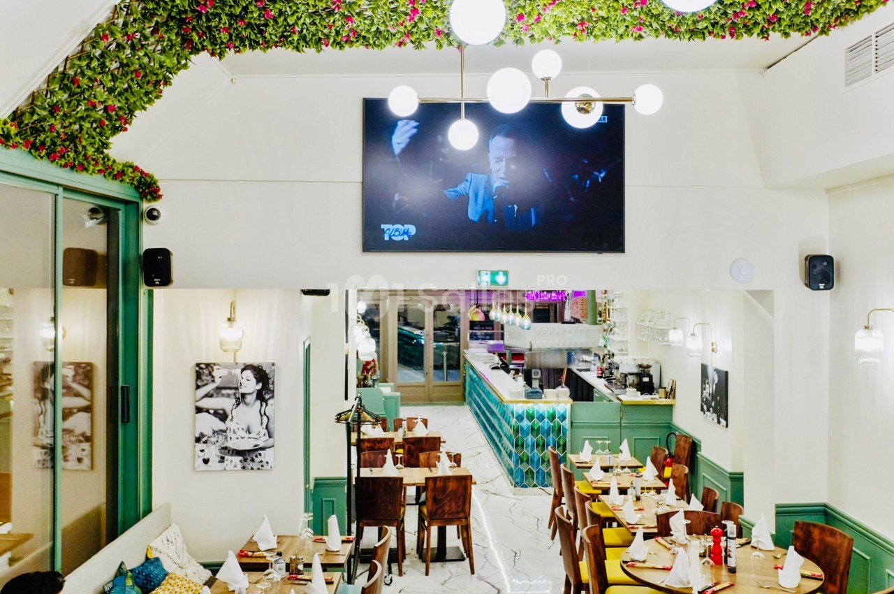 Salle de restaurant lumineuse avec décoration végétale au plafond, tables dressées et écran mural diffusant une vidéo.