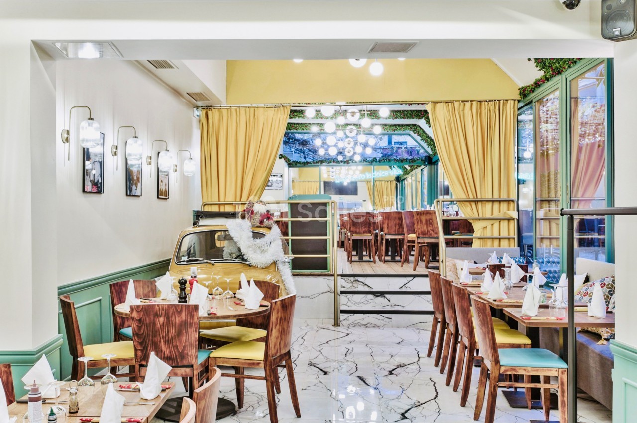 Salle de restaurant lumineuse avec tables dressées, décoration végétale et un espace séparé par des rideaux jaunes.