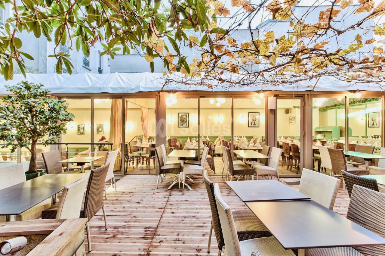 Terrasse en bois aménagée avec des tables et chaises, entourée de végétation et donnant sur une salle lumineuse.