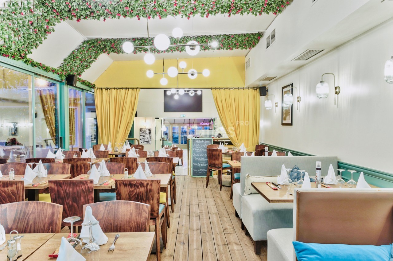 Salle de restaurant lumineuse avec tables dressées, chaises en bois, banquettes et décoration végétale au plafond.