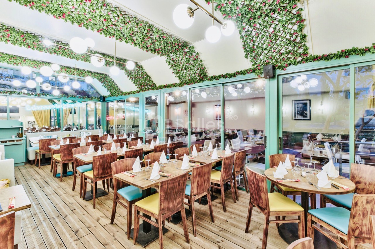 Salle de restaurant lumineuse avec tables en bois dressées, décoration végétale suspendue et grandes baies vitrées.