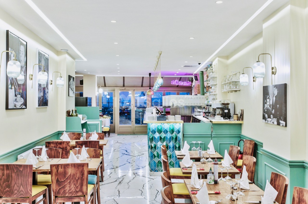 Salle de restaurant lumineuse avec tables dressées, chaises en bois, comptoir moderne et décor pastel.
