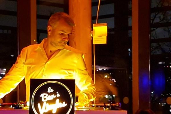 Un homme en chemise blanche se tient derrière une table de mixage éclairée, dans un environnement nocturne.