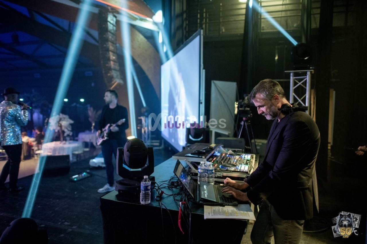 Un DJ mixe sur une scène éclairée, accompagné d'un guitariste et d'un chanteur dans une ambiance de concert.