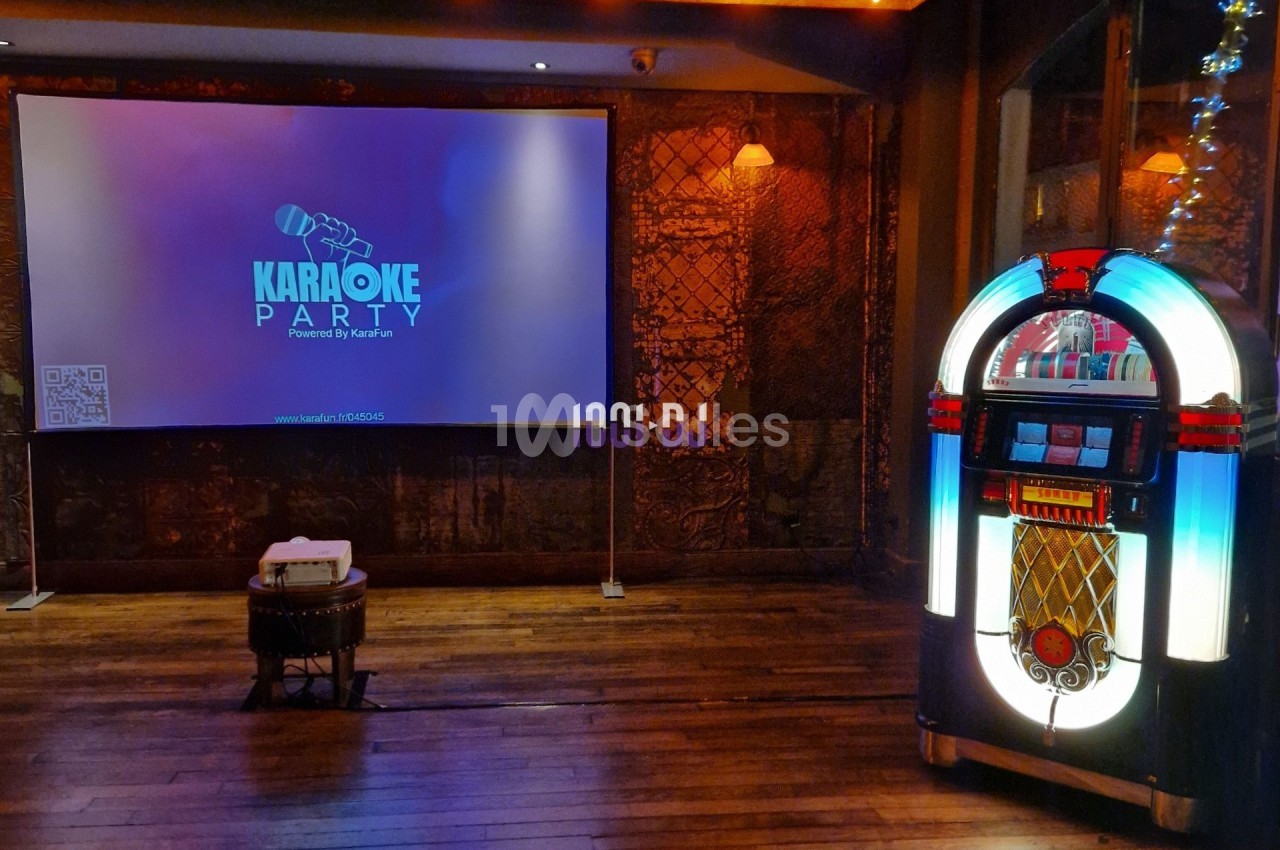 Salle de karaoké avec un jukebox lumineux à droite et un écran de projection affichant ’Karaoke Party’.
