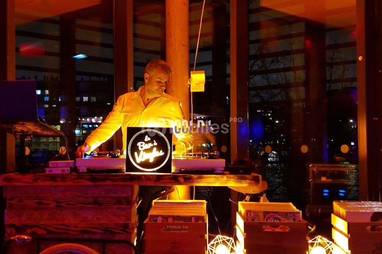 Un DJ mixe de la musique sur un vélo aménagé, entouré de lumières chaleureuses dans un espace vitré en soirée.