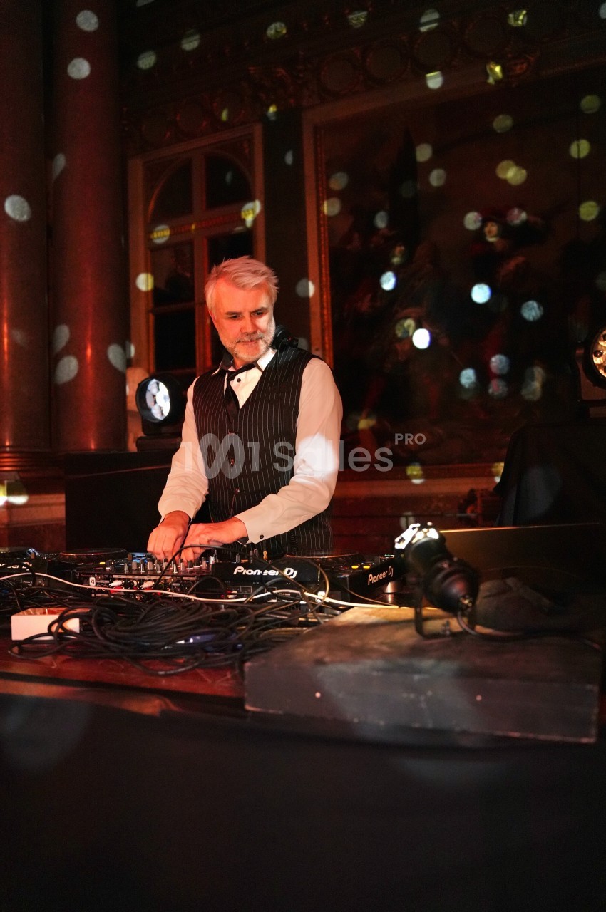 Un homme en costume joue de la musique sur une table de mixage dans une salle éclairée par des lumières colorées.