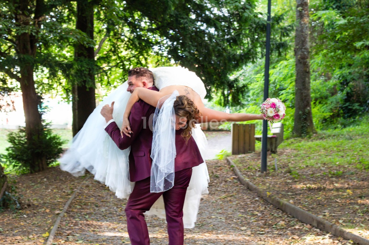 Un marié en costume bordeaux porte sa mariée en robe blanche sur son épaule dans un parc boisé.