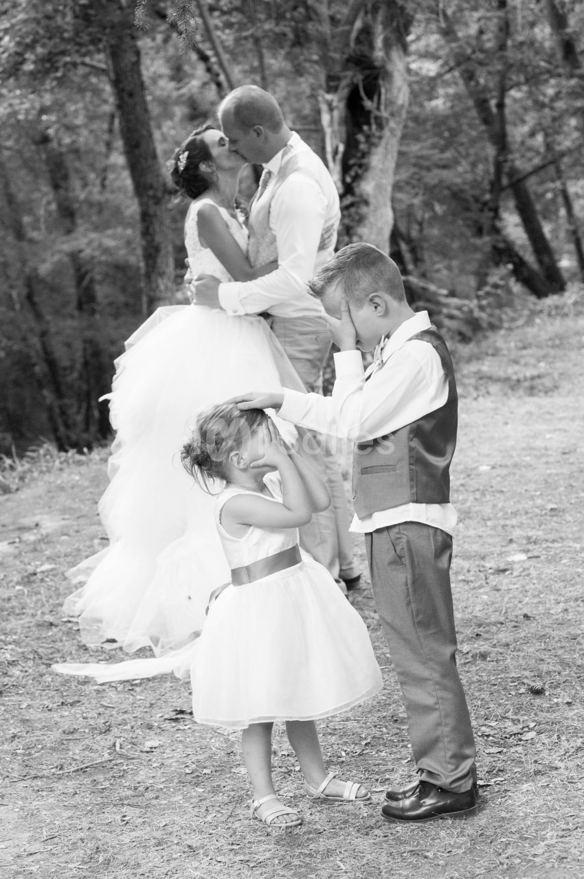 Deux enfants se couvrent les yeux pendant qu'un couple en tenue de mariage s'embrasse dans un cadre boisé.