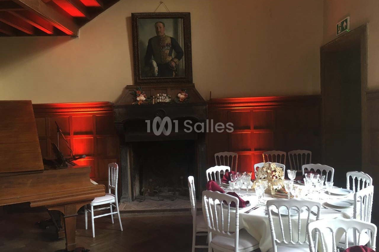 Salle de réception avec cheminée, portrait ancien, piano et tables dressées pour un repas.