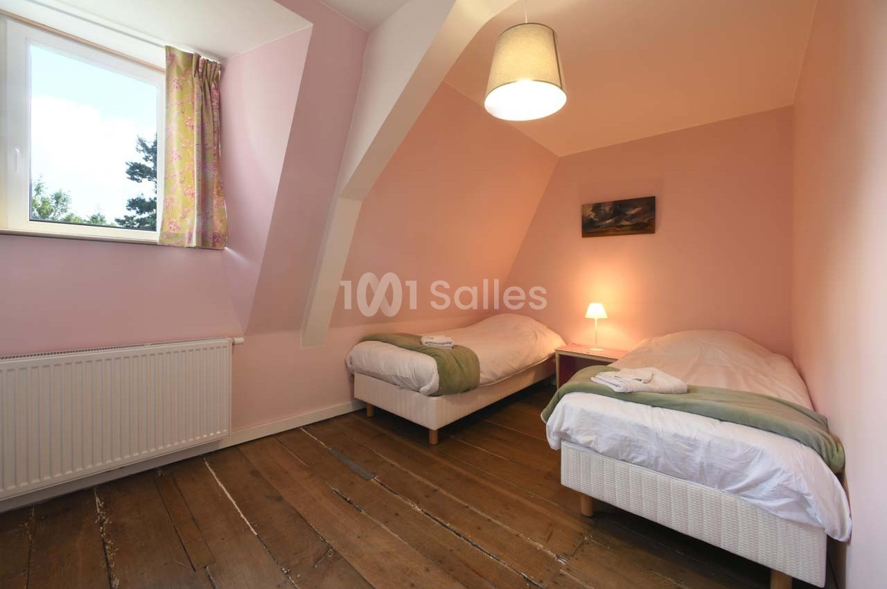 Chambre avec deux lits simples, murs roses, parquet en bois, fenêtre avec rideaux fleuris et lampe allumée.