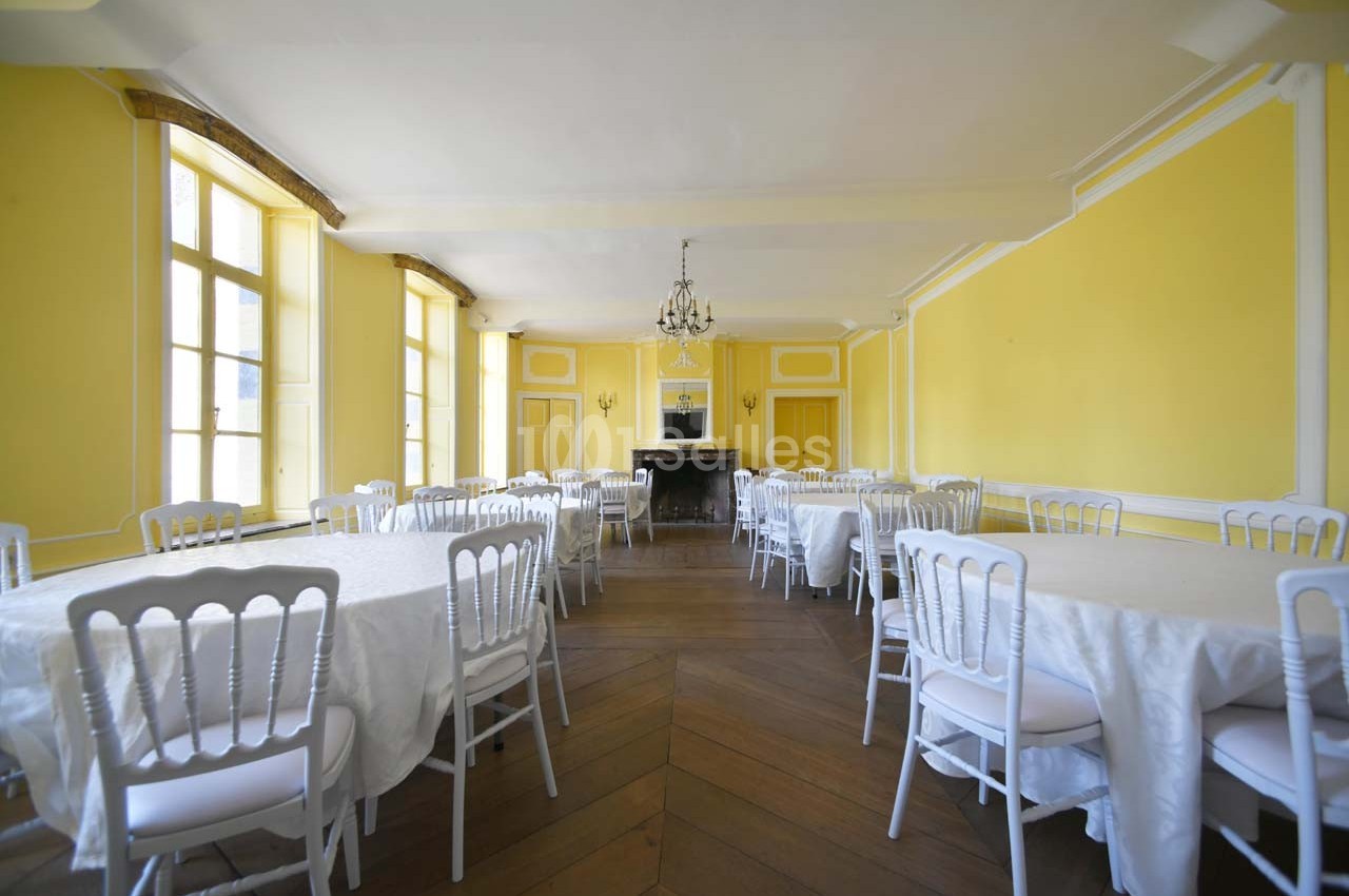 Salle lumineuse avec murs jaunes, parquet en bois, tables rondes dressées de nappes blanches et chaises blanches.