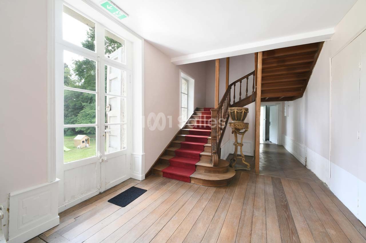 Escalier en bois avec tapis rouge dans une entrée lumineuse, parquet au sol et vue sur un jardin par une porte vitrée.