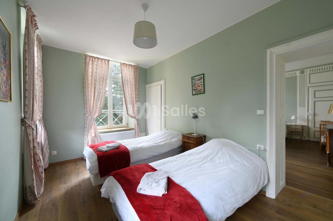 Chambre lumineuse avec deux lits simples, draps blancs, couvertures rouges, parquet et fenêtre avec rideaux roses.