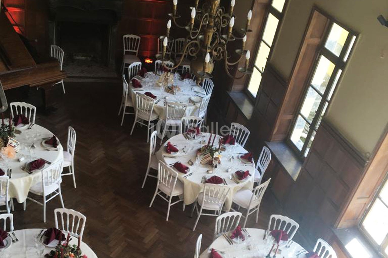Salle de réception élégante avec tables rondes dressées, nappes blanches, serviettes rouges et chandelier central.
