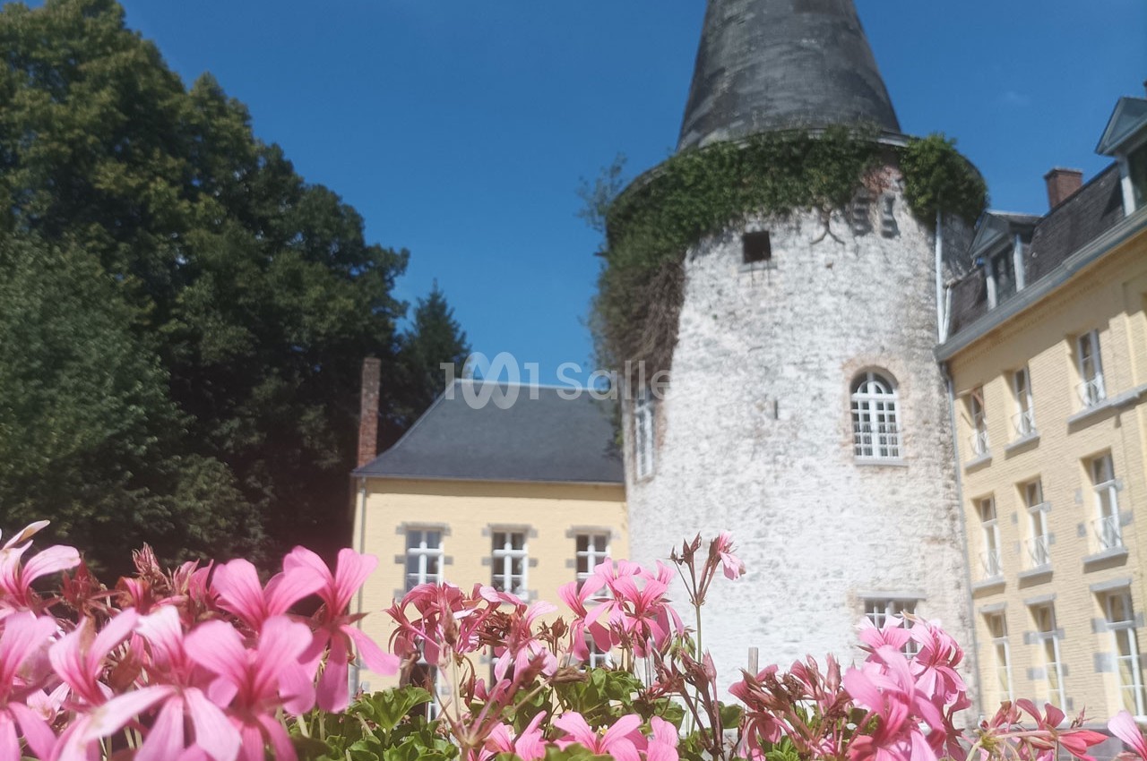 Tour en pierre ancienne recouverte de lierre, entourée de bâtiments jaunes et de géraniums roses au premier plan.