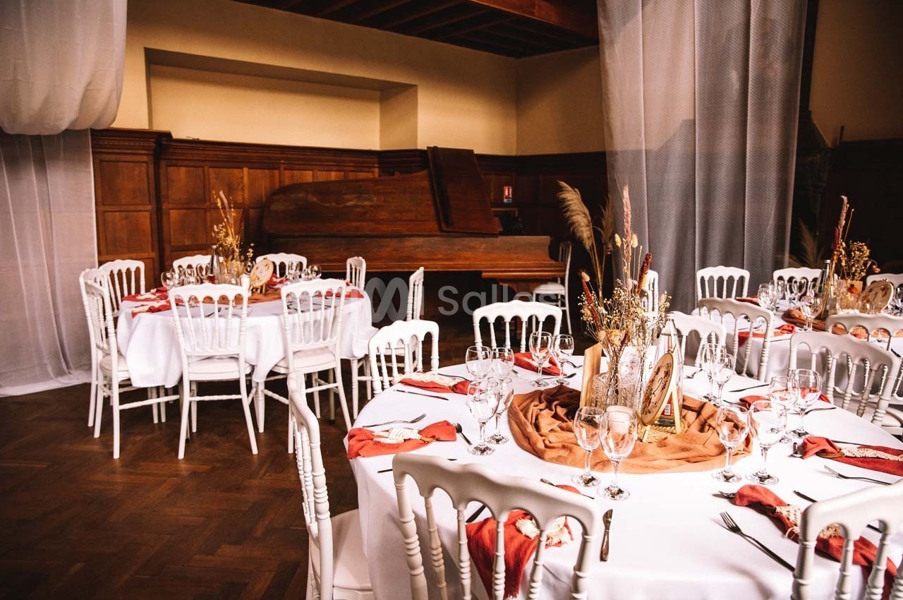 Salle de réception avec tables rondes décorées de nappes blanches, serviettes rouges et centres de table floraux.