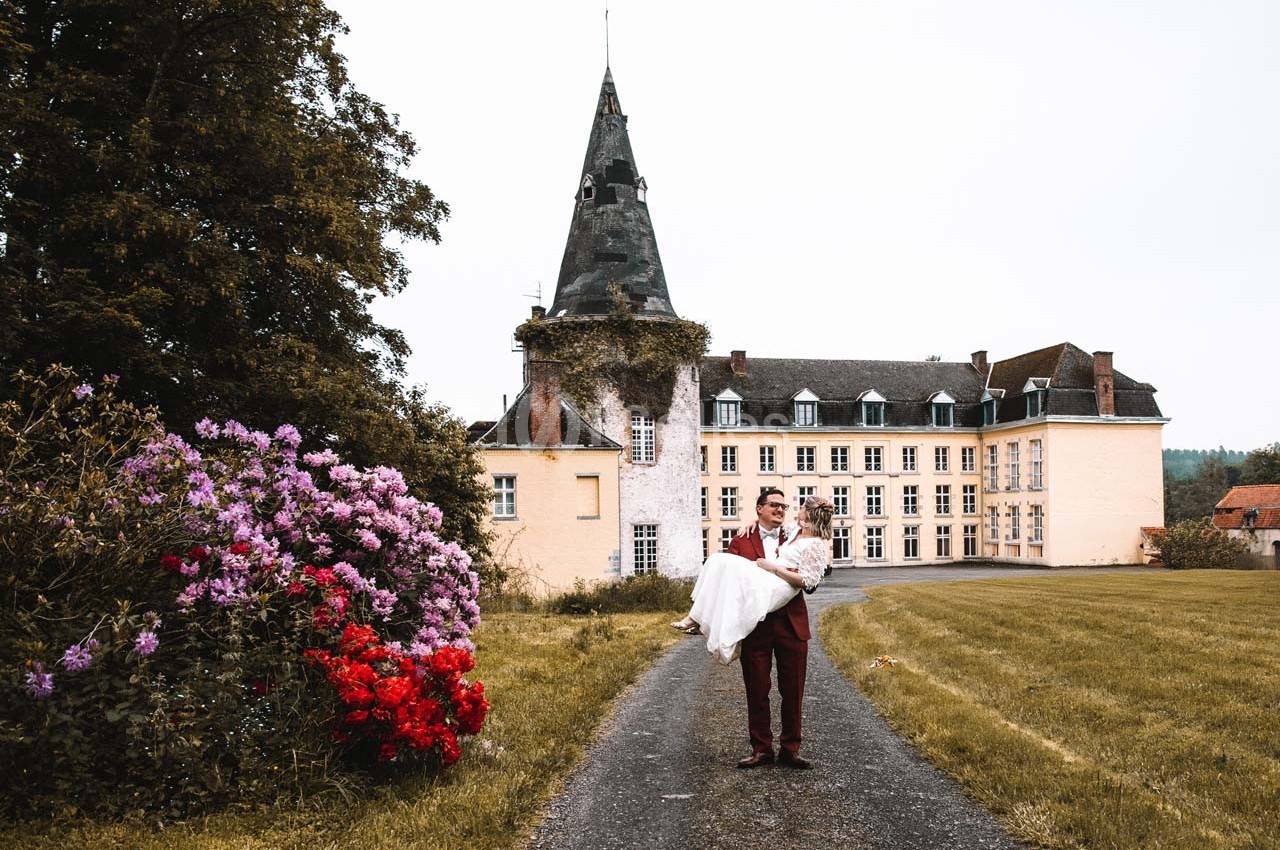 Un homme en costume porte une femme en robe de mariée devant un château entouré de verdure et de fleurs.