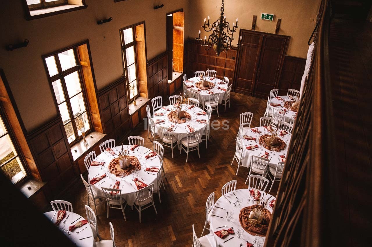 Salle de réception avec tables rondes dressées, nappes blanches, serviettes rouges et lumière naturelle entrant par les…