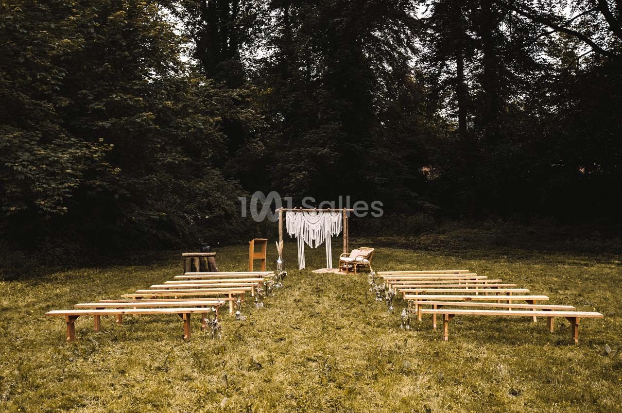 Allée centrale bordée de bancs en bois menant à une arche décorée de tissu blanc dans un cadre naturel boisé.