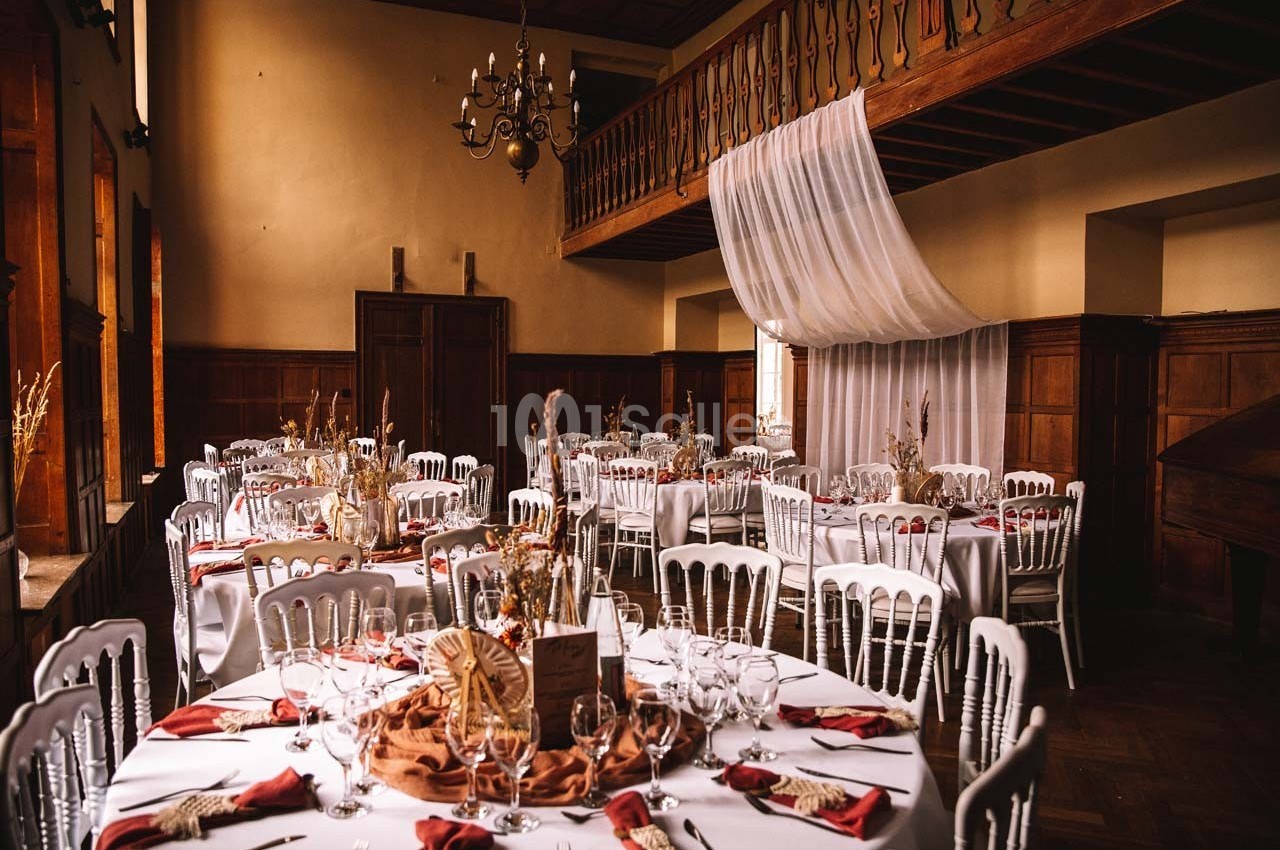 Salle de réception avec tables rondes dressées, nappes blanches, serviettes rouges et décoration champêtre.