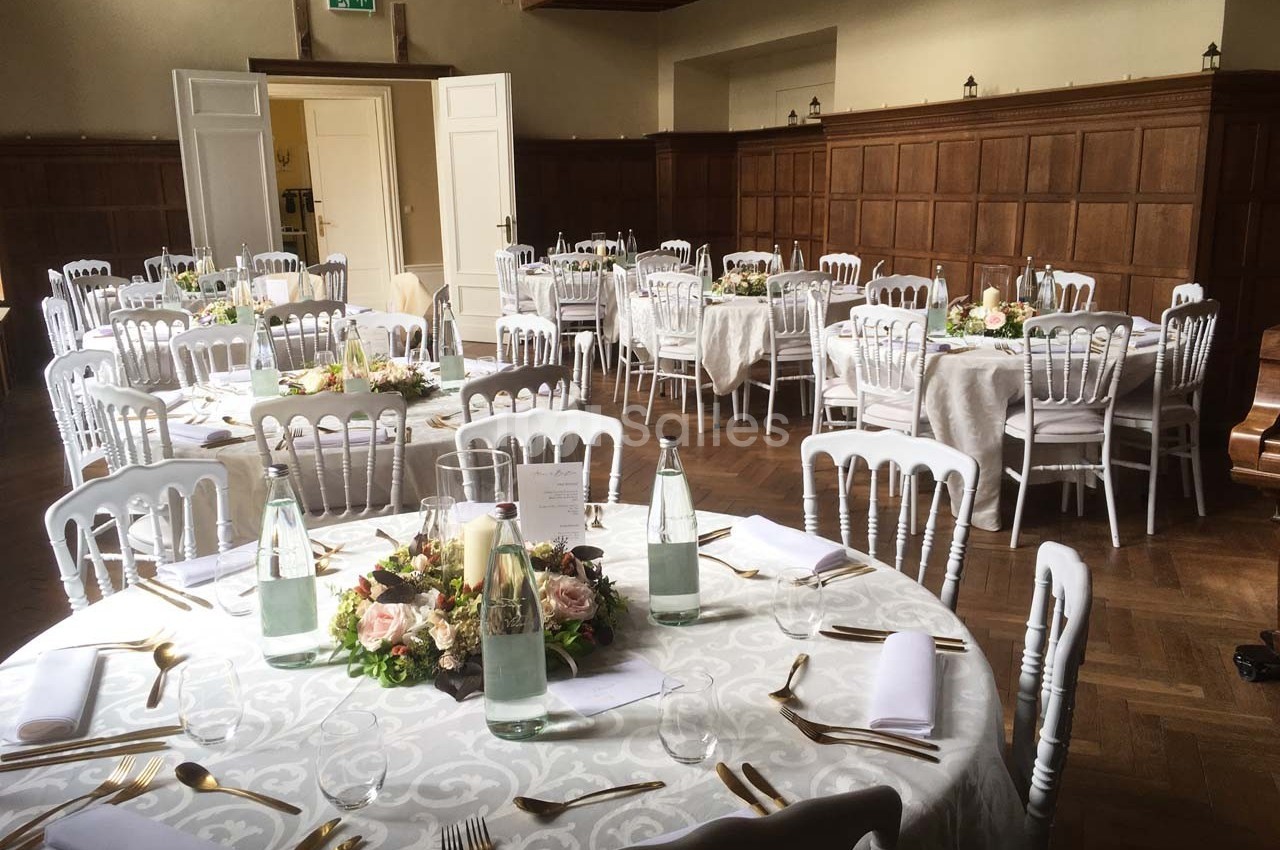 Salle de réception avec tables rondes dressées, nappes blanches, chaises blanches et décorations florales au centre.