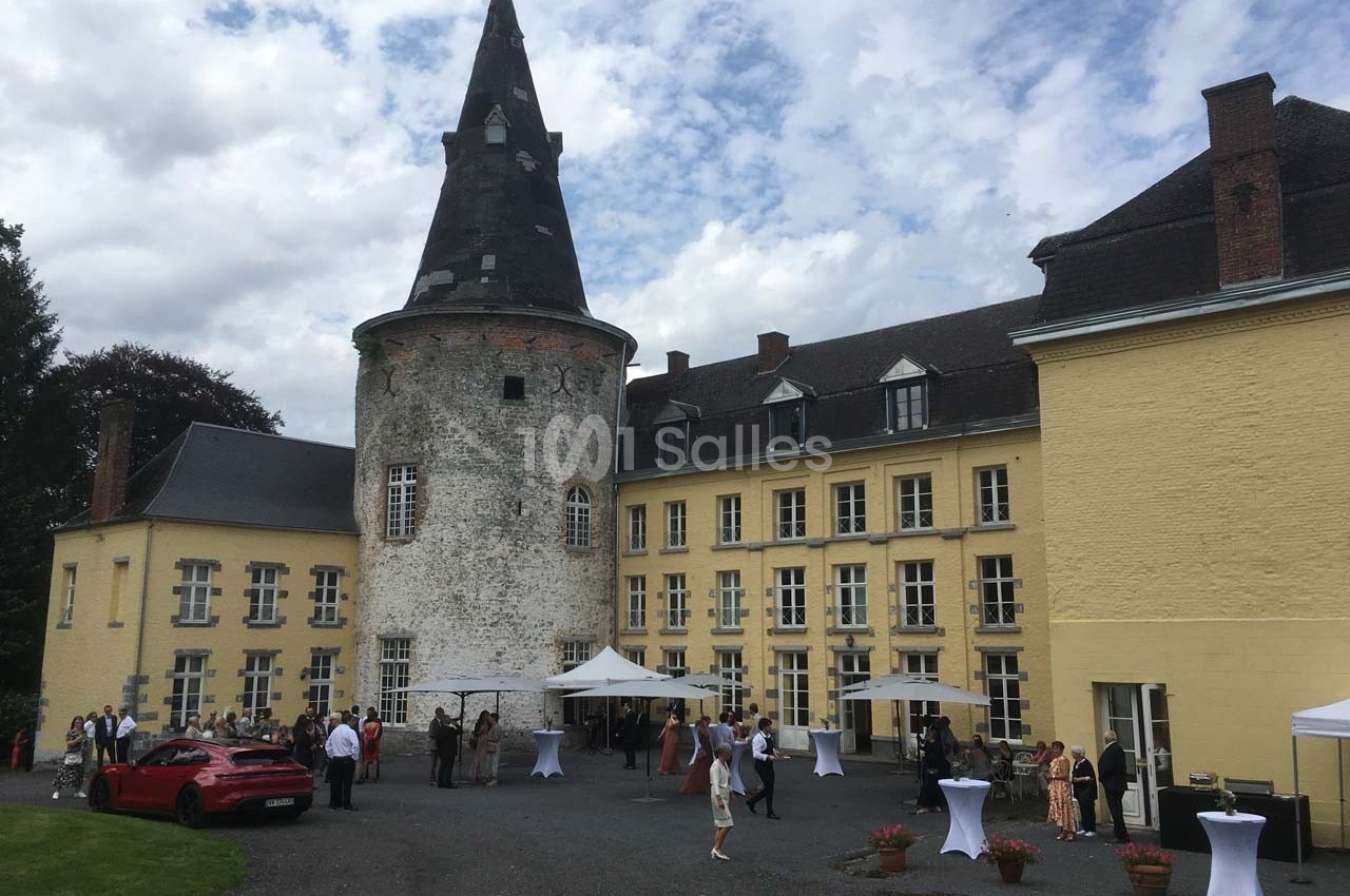 Château avec tour en pierre, façade jaune et invités rassemblés autour de tables en extérieur par temps nuageux.