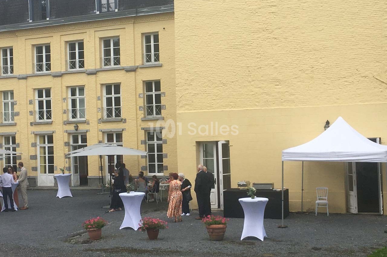 Personnes rassemblées dans une cour pavée devant un bâtiment jaune, avec des tables hautes et des tentes blanches.