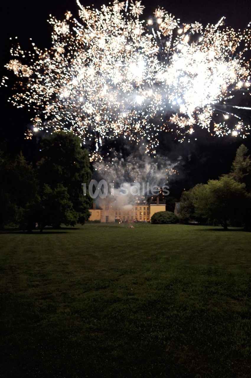 Feu d'artifice illuminant le ciel nocturne au-dessus d'un grand bâtiment entouré de pelouses et d'arbres.
