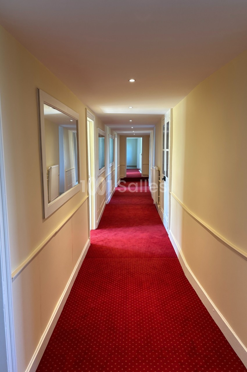Couloir étroit avec moquette rouge, murs clairs, miroir mural et portes alignées des deux côtés.