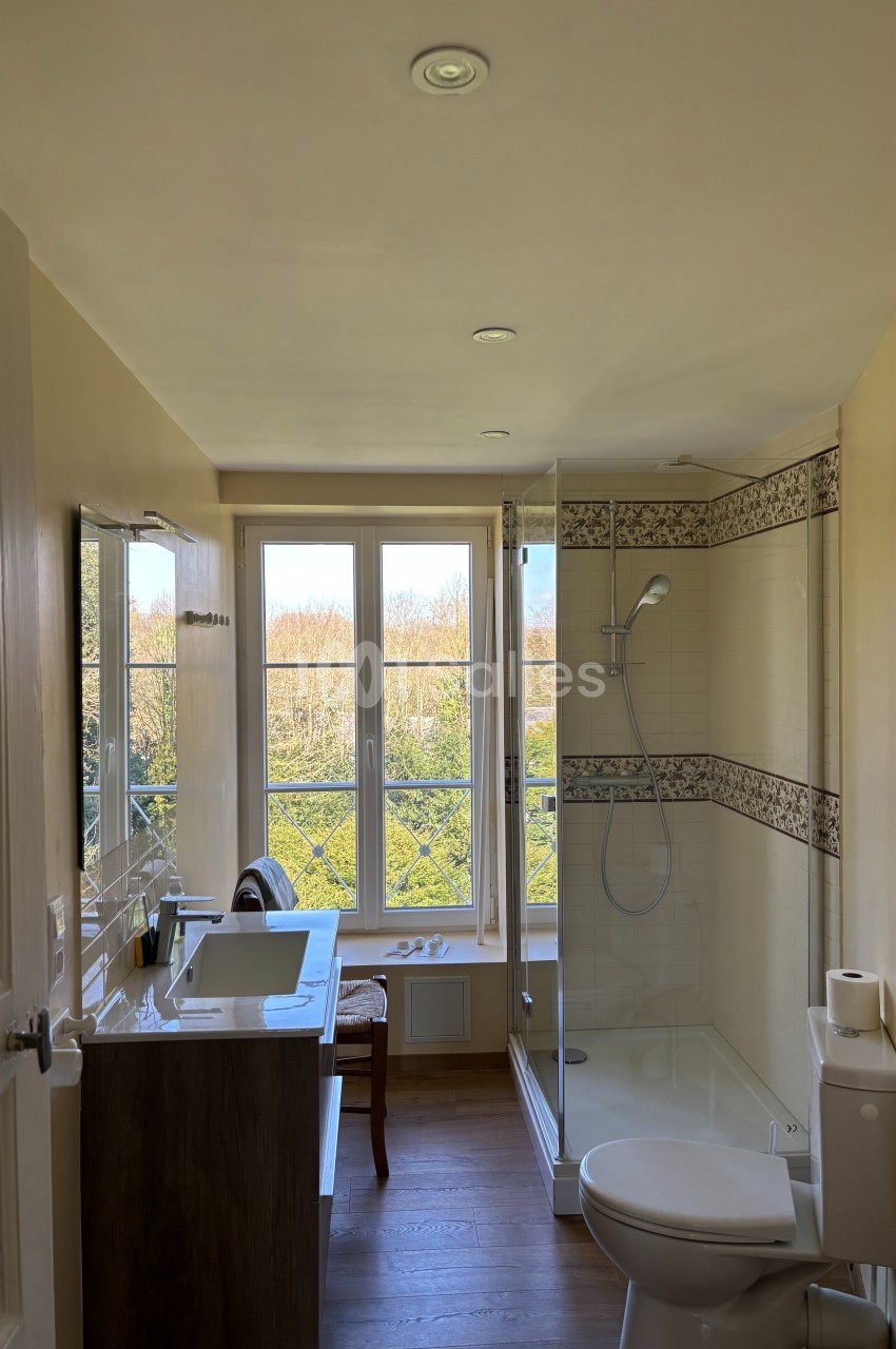Salle de bain lumineuse avec douche vitrée, lavabo, toilettes et grande fenêtre donnant sur un paysage arboré.