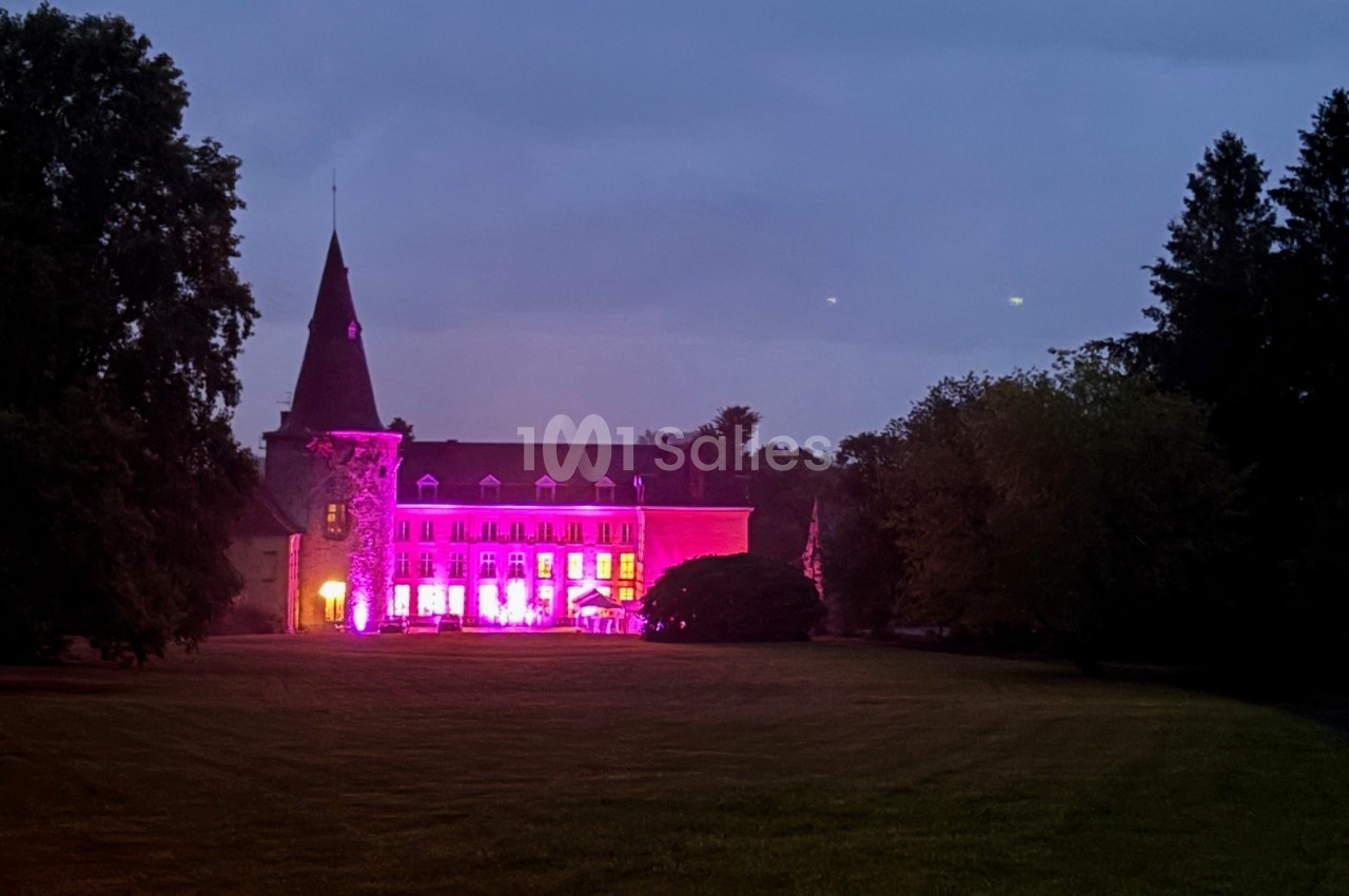 Façade d'un bâtiment ancien éclairé par des lumières roses et violettes, entouré d'arbres au crépuscule.