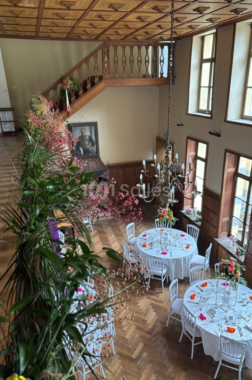 Salle de réception élégante avec tables rondes décorées de fleurs, parquet en bois et escalier en arrière-plan.