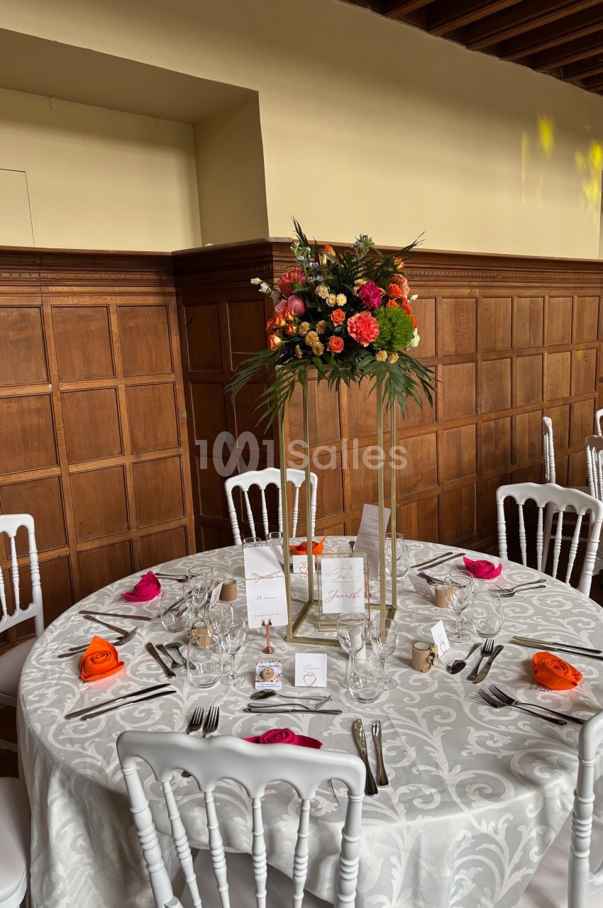 Table ronde décorée pour un événement, avec nappe blanche, chaises blanches et centre de table floral coloré.