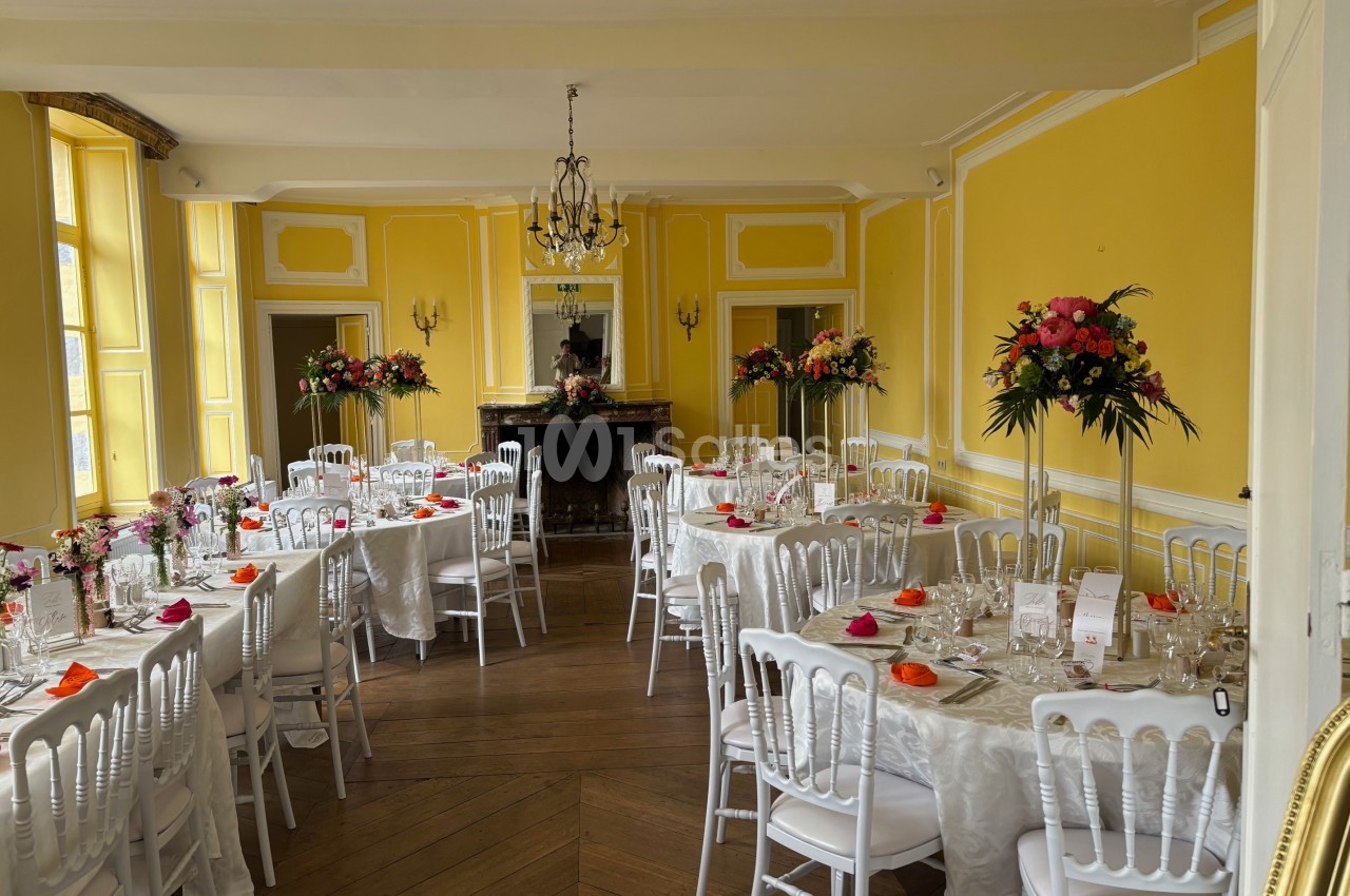 Salle de réception lumineuse avec murs jaunes, tables dressées, chaises blanches et décorations florales colorées.