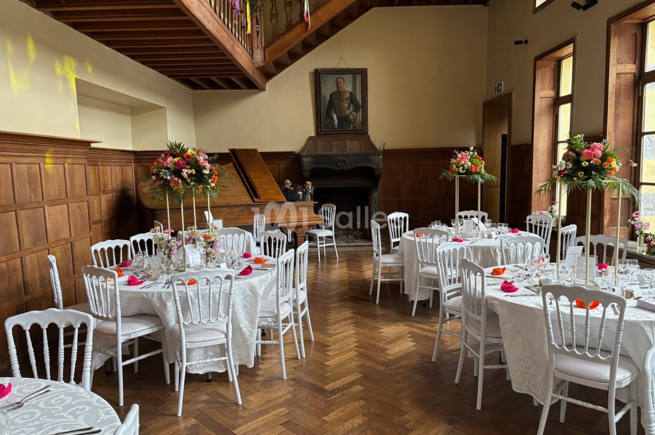 Salle de réception élégante avec tables dressées, chaises blanches, décorations florales et un piano en arrière-plan.
