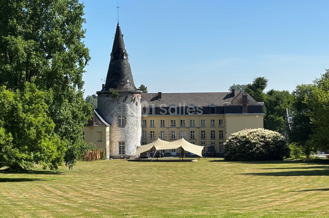Château avec tour en pierre, pelouse entretenue et tente blanche dans un cadre verdoyant sous un ciel bleu.