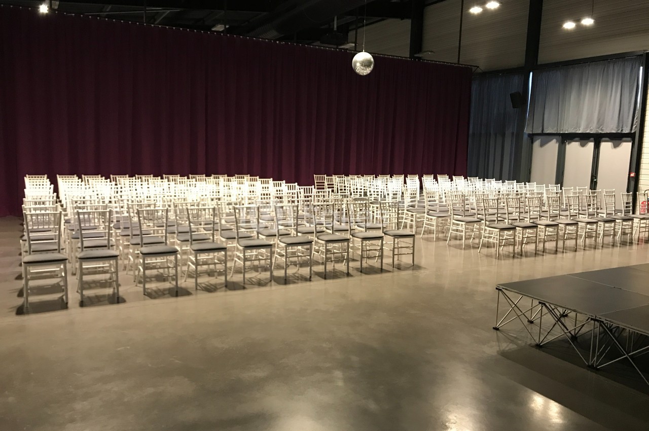 Salle de conférence avec des rangées de chaises blanches alignées devant une scène et un rideau violet.
