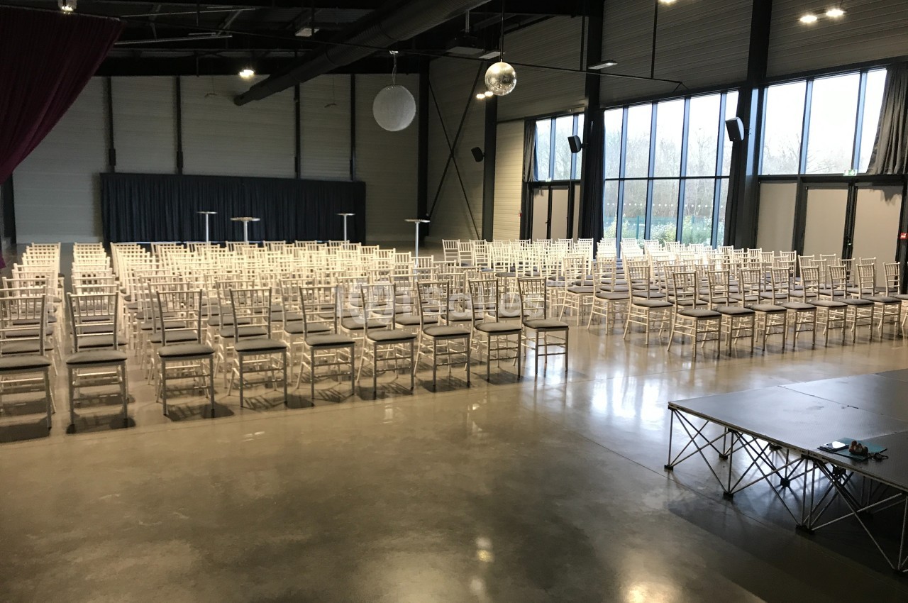Salle de réception avec rangées de chaises blanches alignées, scène à droite et grandes baies vitrées en arrière-plan.