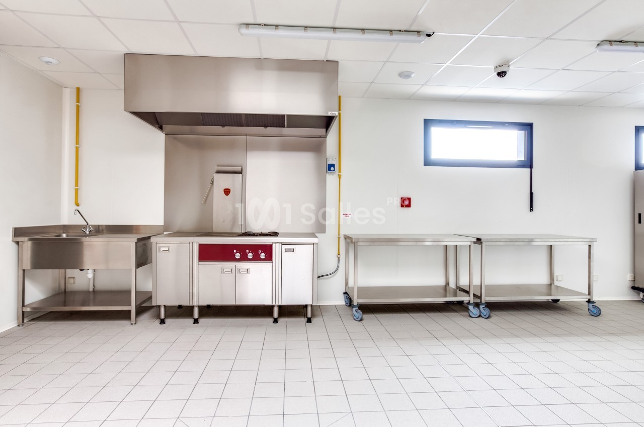 Cuisine professionnelle avec évier, hotte en inox, plan de travail sur roulettes et carrelage au sol.
