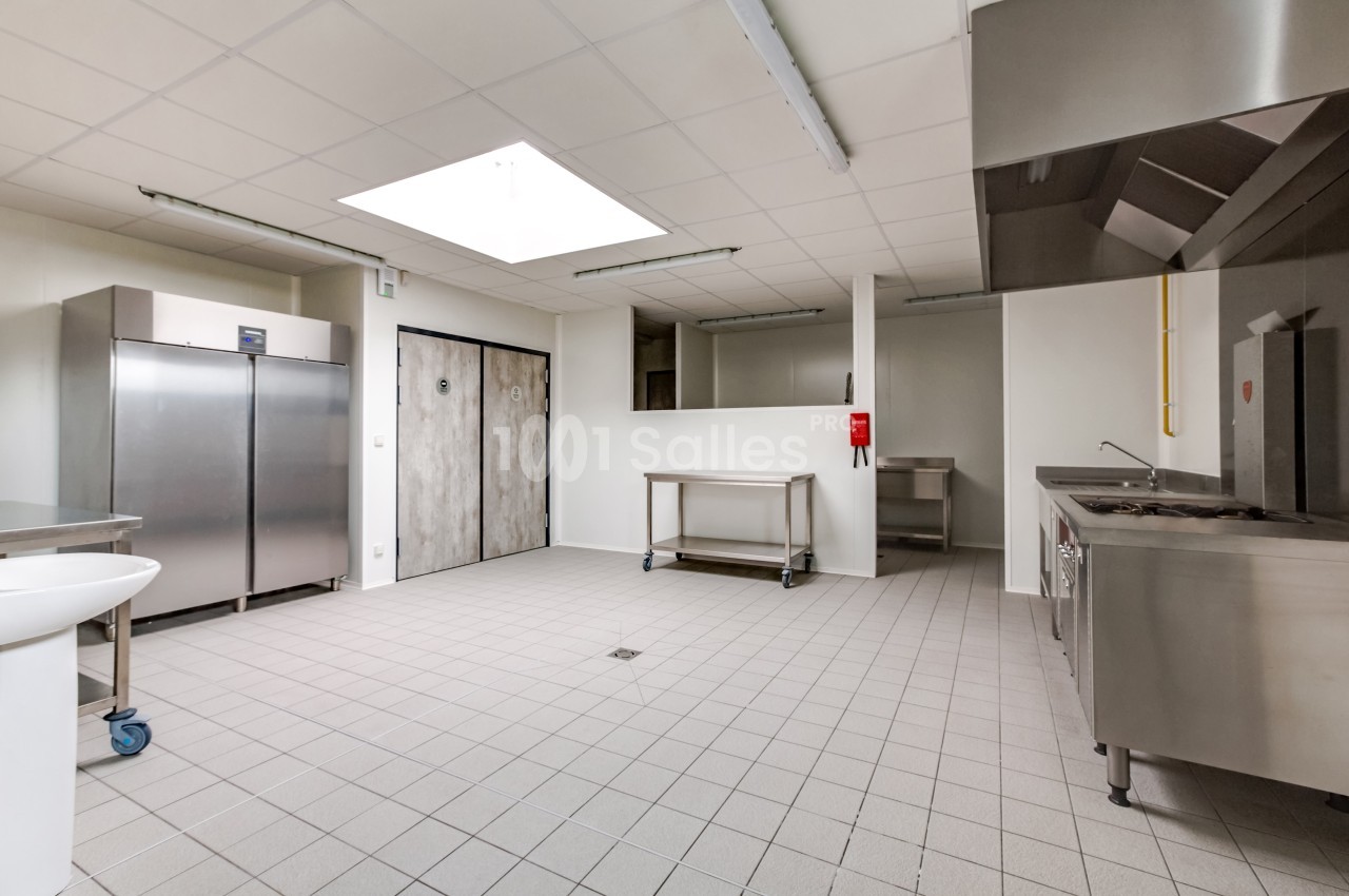Cuisine professionnelle vide avec équipements en inox, sol carrelé, éclairage naturel et portes de stockage visibles.