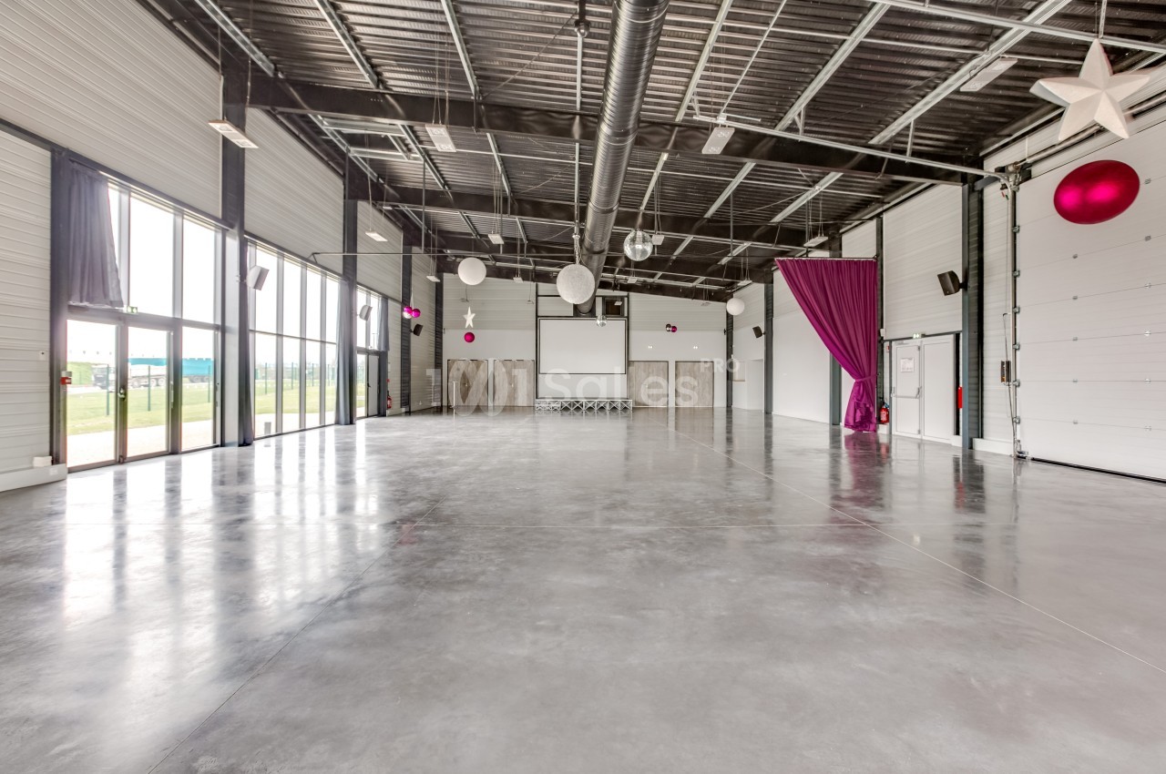 Salle spacieuse avec sol en béton, grandes baies vitrées, rideau violet, décorations suspendues et plafond industriel.