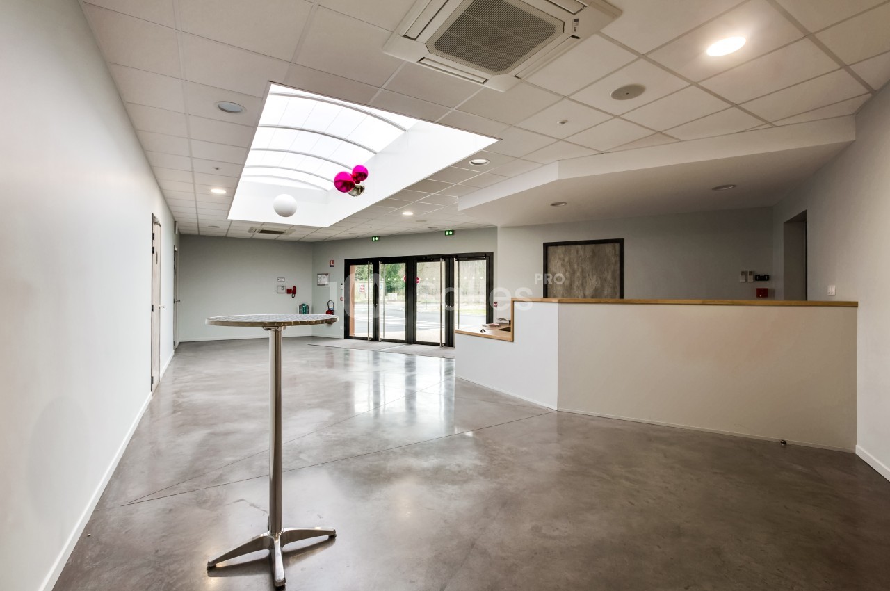 Salle lumineuse avec sol en béton, plafond à dalles blanches, grande baie vitrée et table haute en métal.