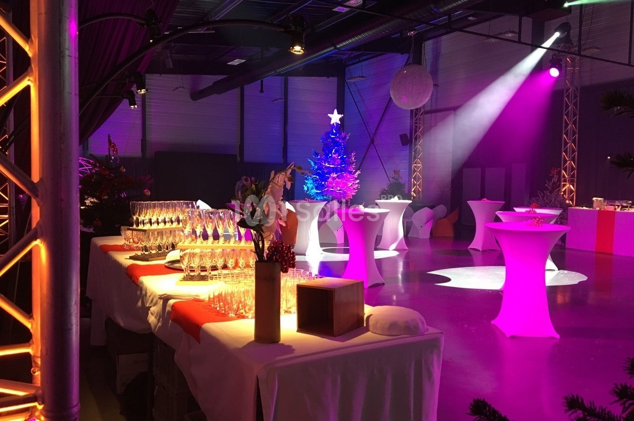Salle décorée pour un événement festif avec tables hautes, éclairages colorés et sapin illuminé.