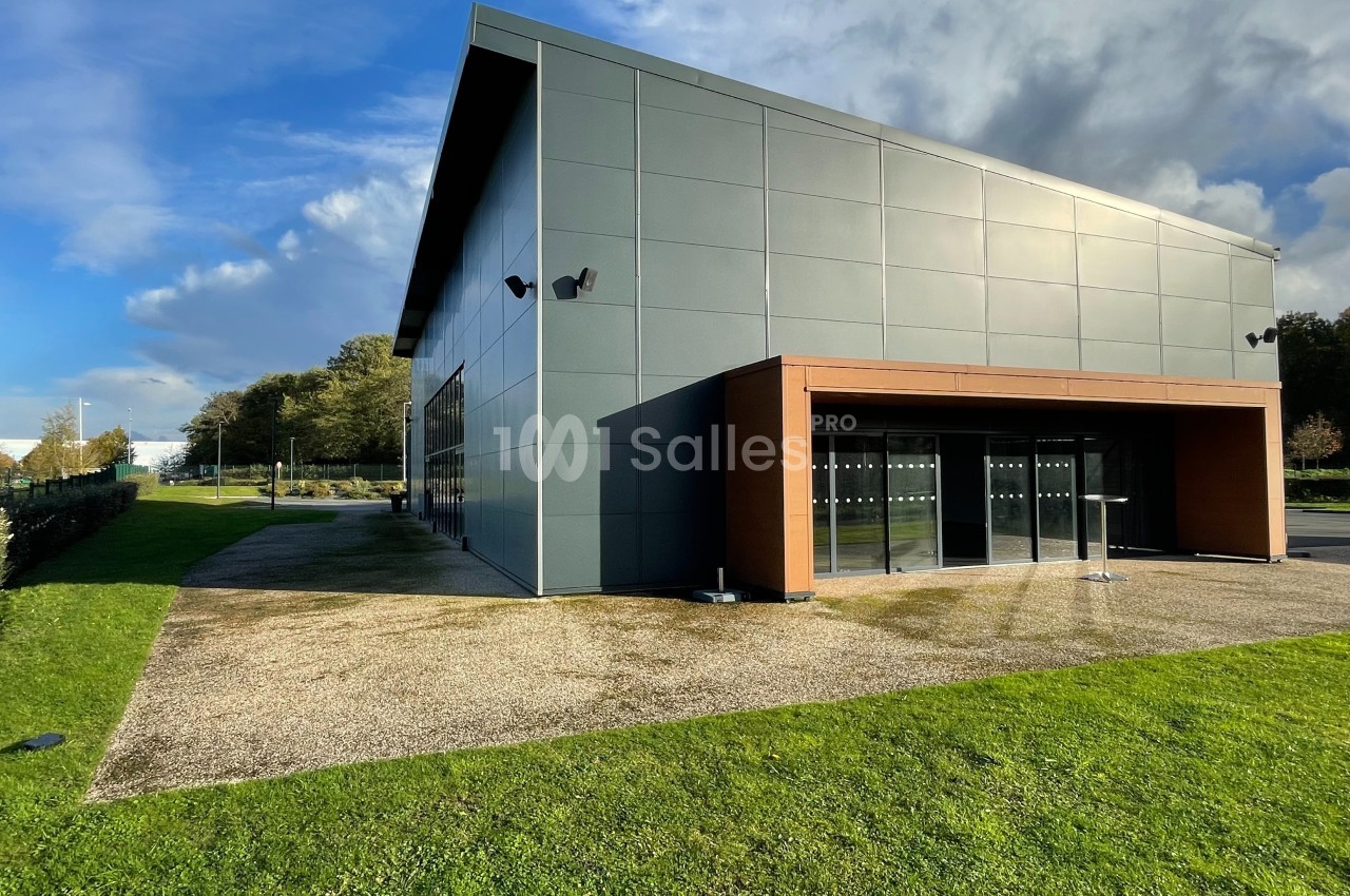 Bâtiment moderne avec façade en panneaux gris, grandes baies vitrées et espace vert au premier plan.
