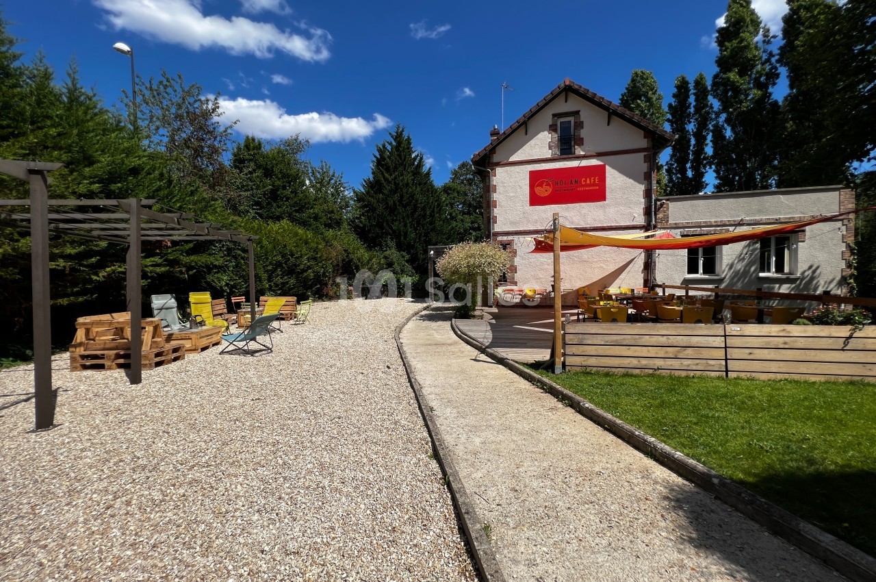 Cour extérieure avec terrasse aménagée, tables, chaises et pergola, entourée de verdure et d'un bâtiment en arrière-plan.