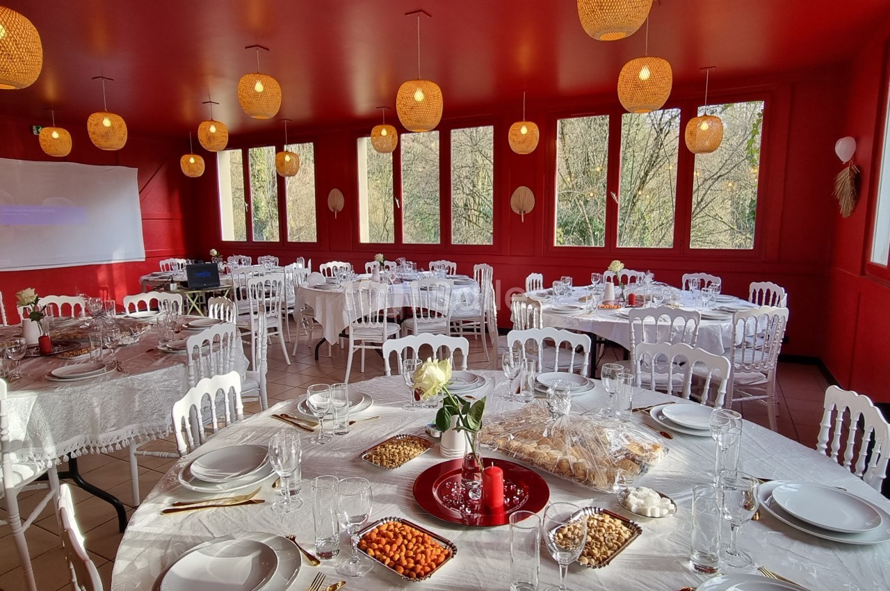 Salle de réception lumineuse avec murs rouges, tables rondes dressées de nappes blanches et décorations suspendues.