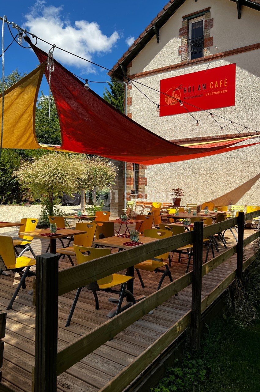 Terrasse en bois avec des tables et chaises jaunes, ombragée par des voiles colorées, devant un bâtiment clair.