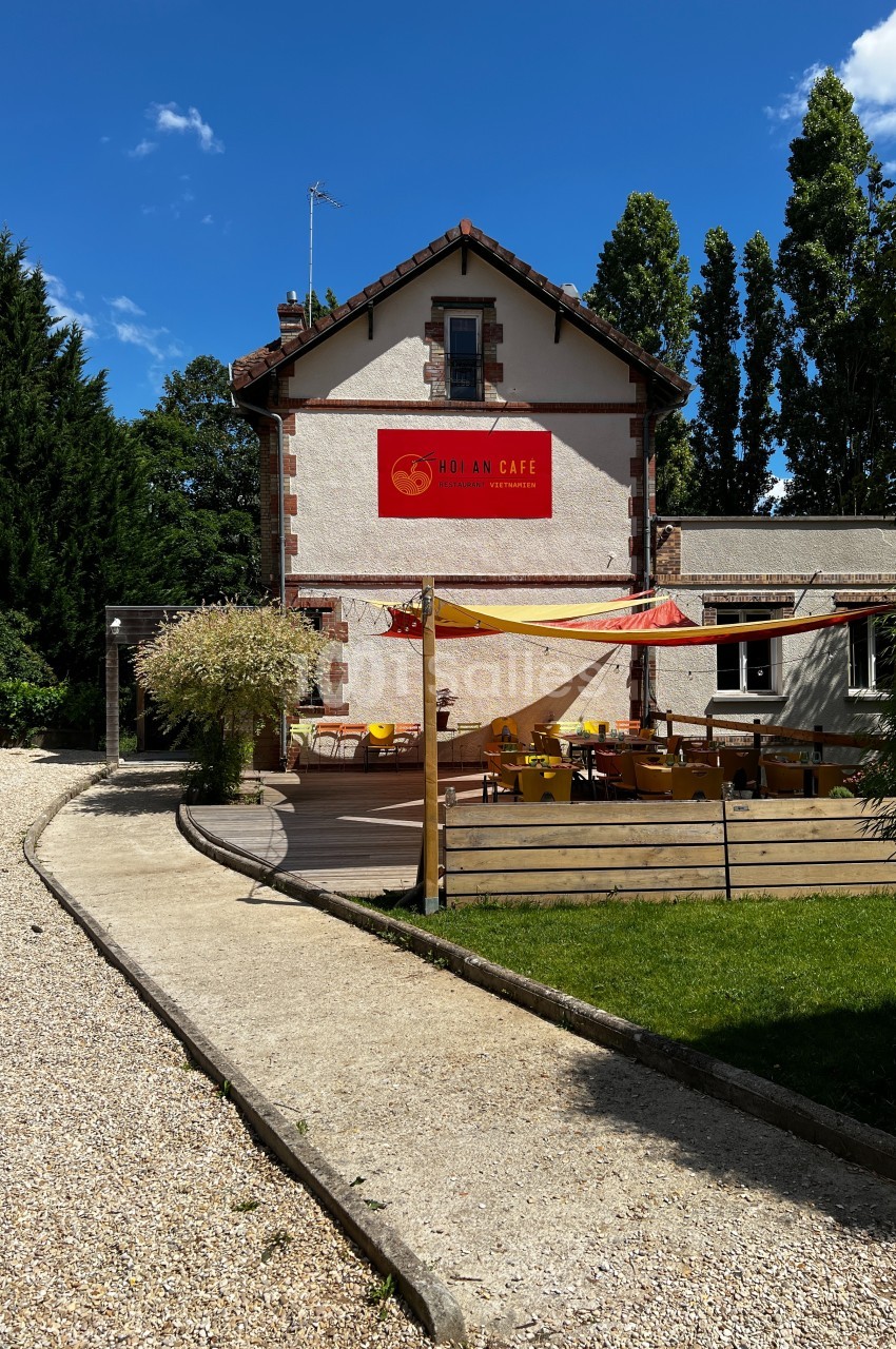 Façade d'un café avec terrasse extérieure aménagée, entourée de végétation sous un ciel bleu.